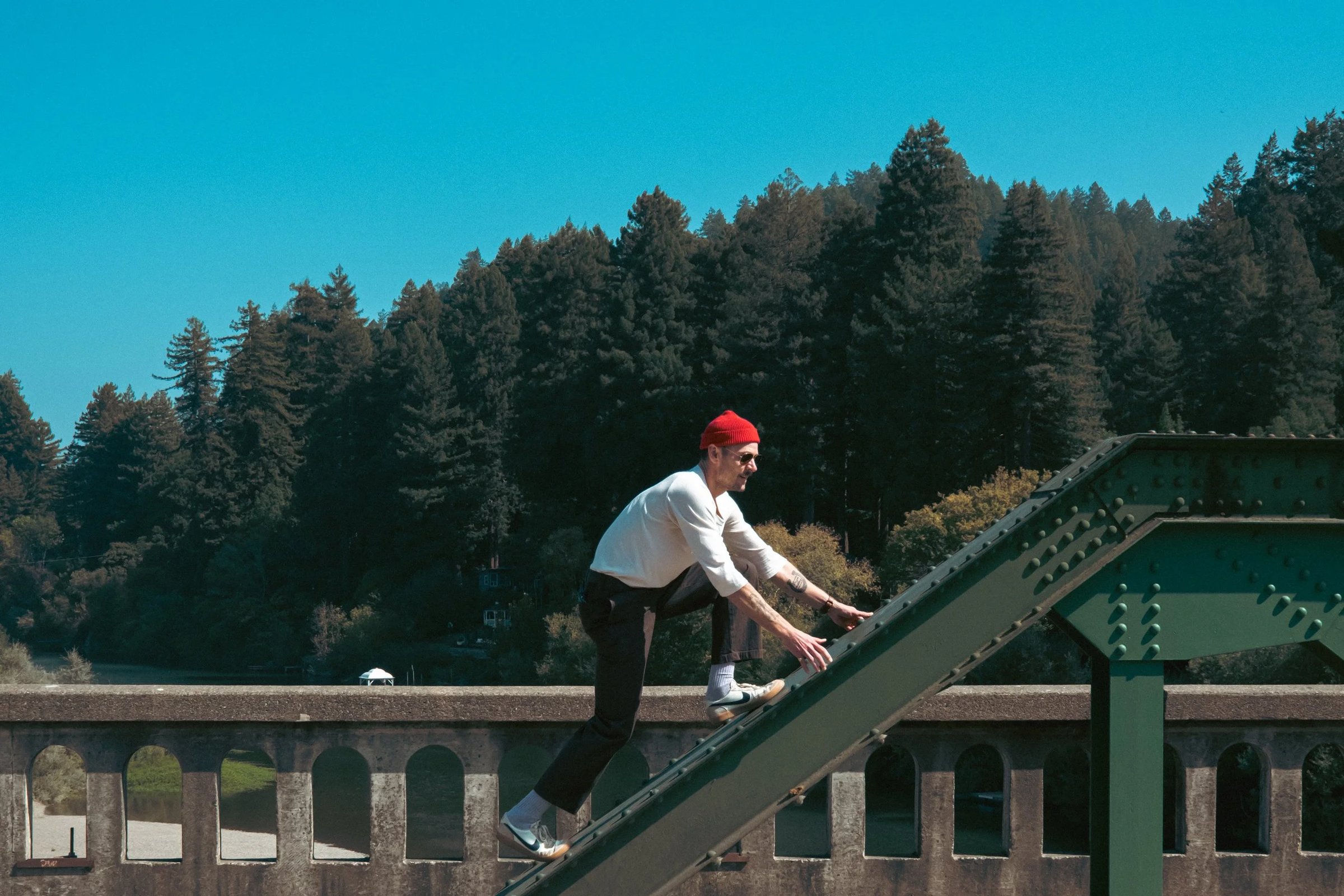 A person in a red beanie and white shirt climbs a green steel structure against a backdrop of tall trees and blue sky.