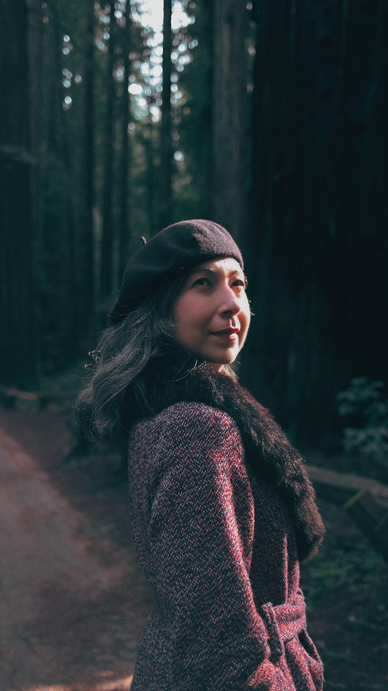 A woman in a beret and coat stands in a redwood forest, illuminated by soft sunlight filtering through the trees.