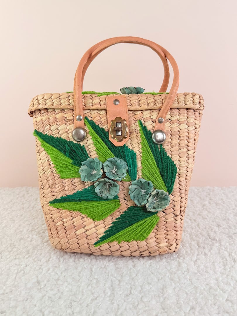 Woven straw handbag with blue floral and green leaf embellishments, featuring a tan leather handle and clasp closure, inspired by 1970s design.