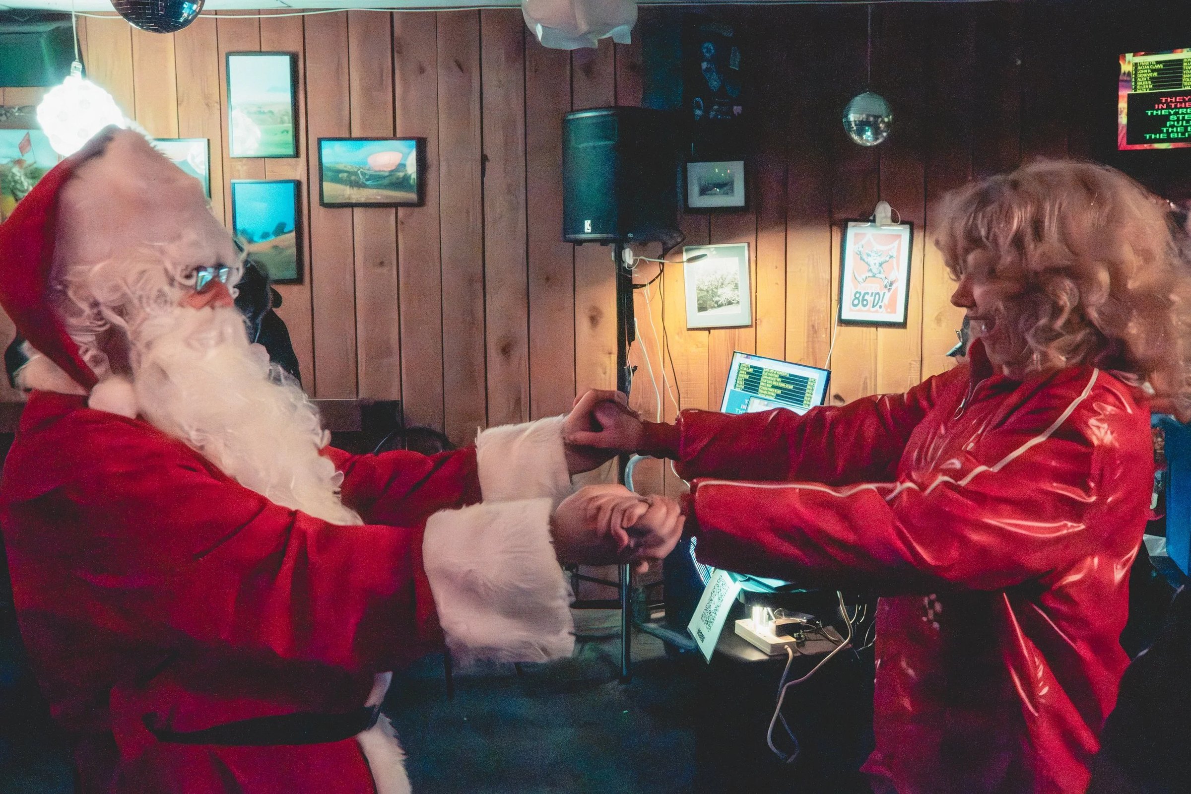 Two individuals dressed in Santa costumes are holding hands in a room with wooden walls and various electronic equipment.