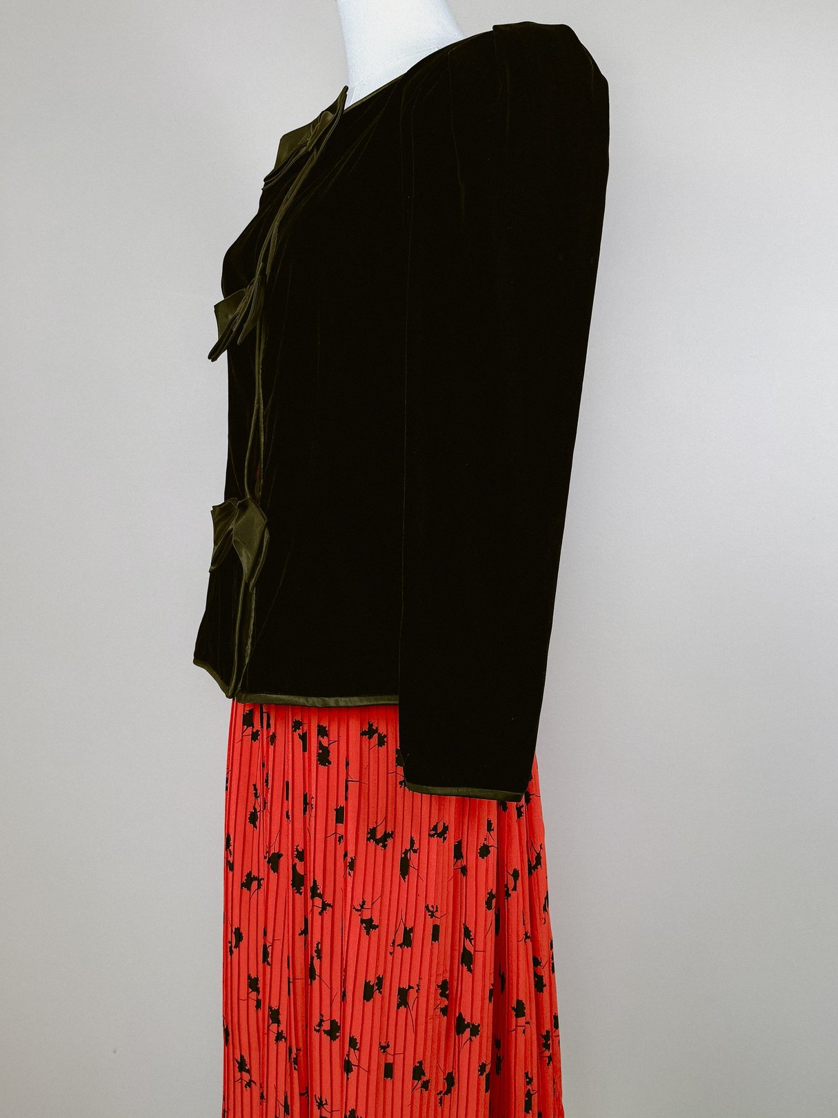 The black velvet jacket features a structured silhouette, long sleeves, and bow detailing, styled with a red floral pleated skirt.