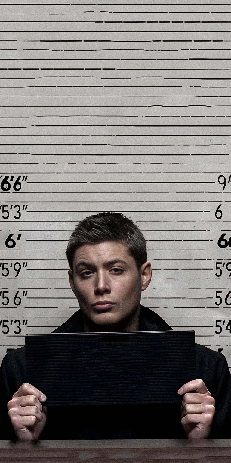 A classic 4k wallpaper featuring Dean Winchester (Jensen Ackles) from the long-running series Supernatural. This image recreates the iconic police station mugshot scene, showing Dean holding the booking board against a height chart background. Essential for any member of the SPN Family.