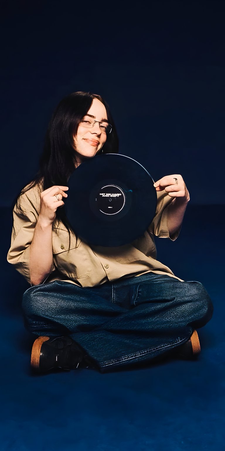 Download this stunning Billie Eilish 4K wallpaper for your iPhone, featuring the singer holding a vinyl record. The image showcases her wearing glasses, and dark blue jeans, all set against a dark blue background.