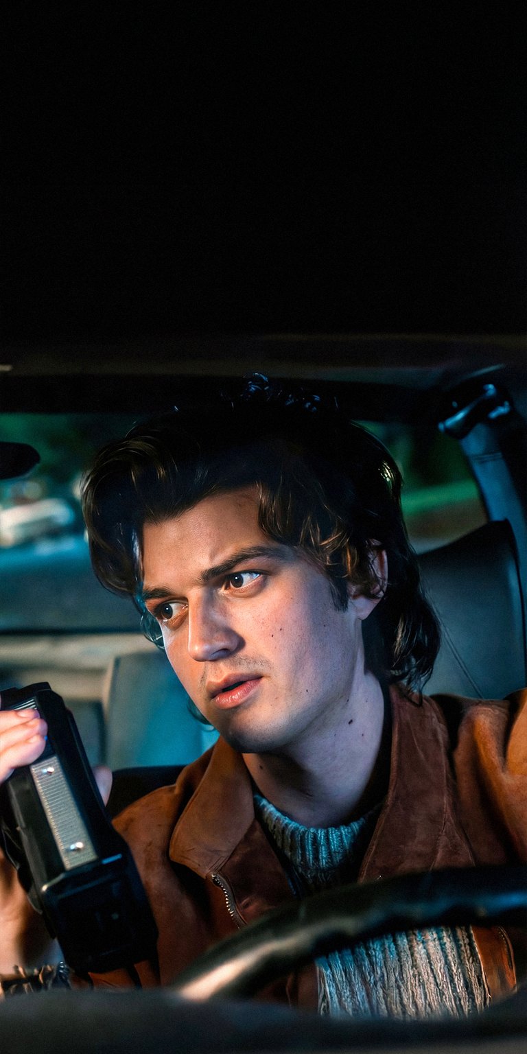 Download this captivating Steve Harrington 4K wallpaper for your iPhone, showcasing his intense expression and retro camcorder. The warm brown suede jacket and vintage blue sweater complete the nostalgic vibe of this stunning wallpaper.