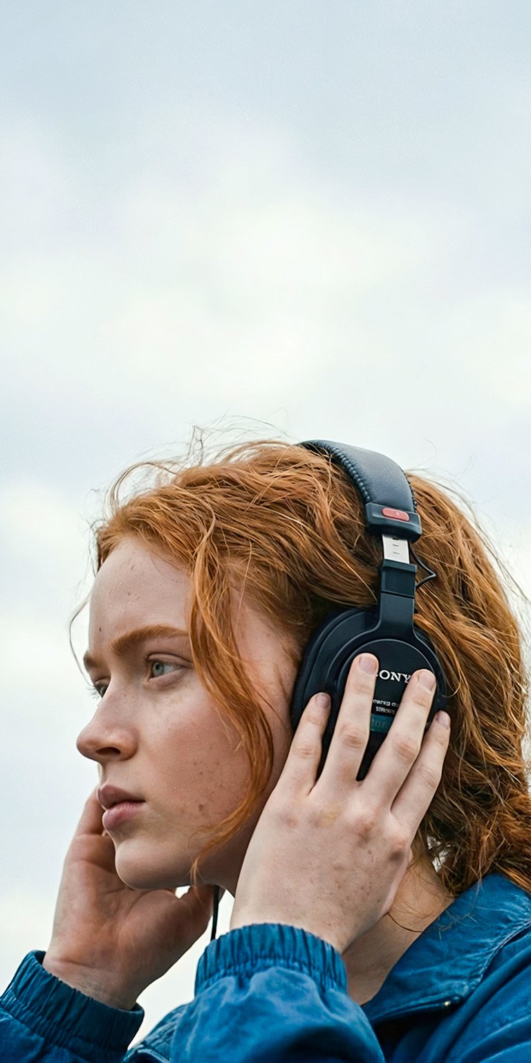 A striking, contemplative 4K portrait of Max Mayfield (Sadie Sink) with her signature red hair and headphones. The composition is clean and atmospheric, featuring her against a vast, bright, overcast sky. This high-resolution image highlights her pensive expression, making it a beautiful and introspective background for fans of the character.