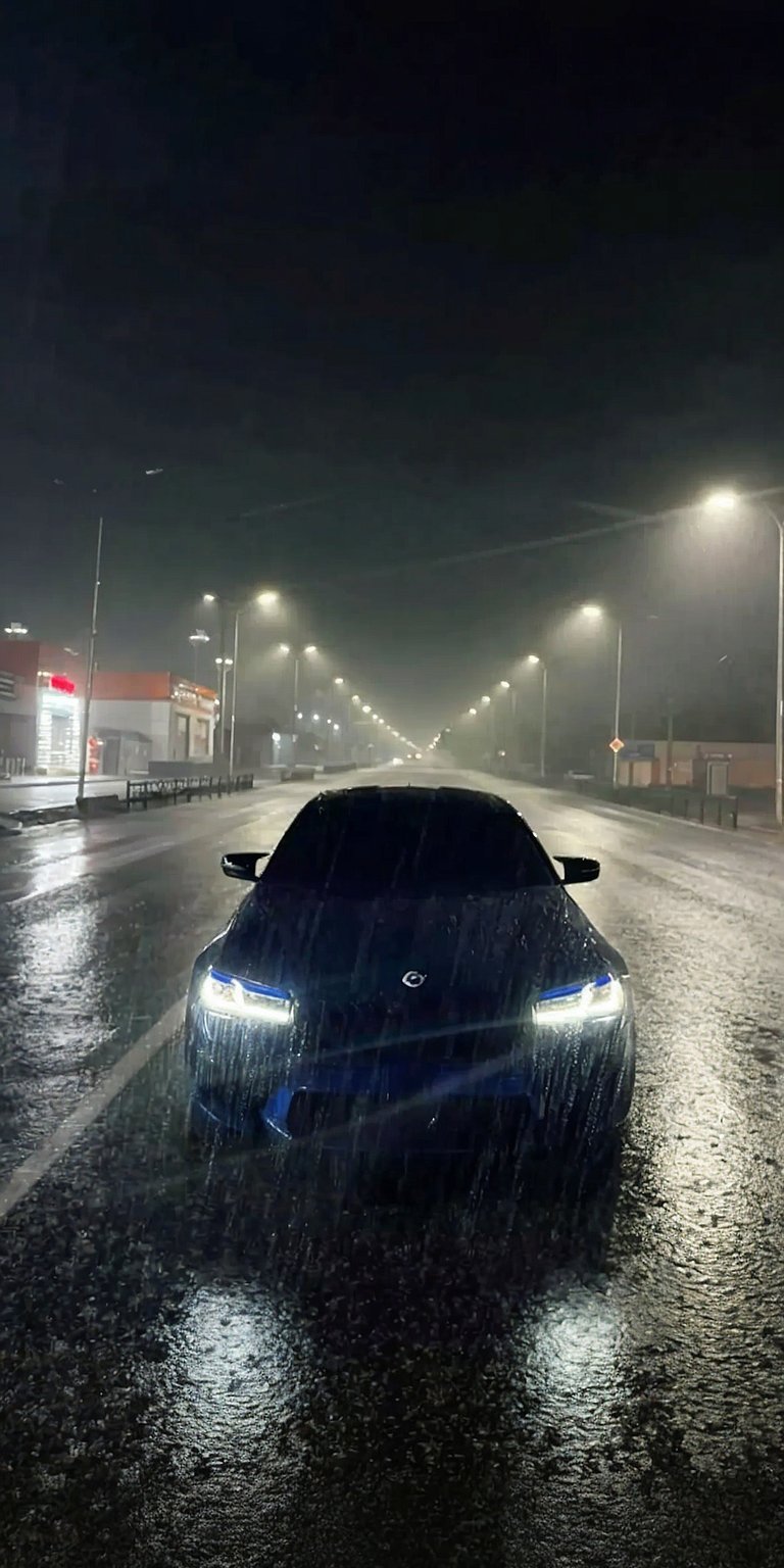 Experience the sleek BMW M4 in stunning 4K resolution with this captivating wallpaper for your iPhone. See the striking headlights illuminate the rain-slicked road under the glow of streetlights.