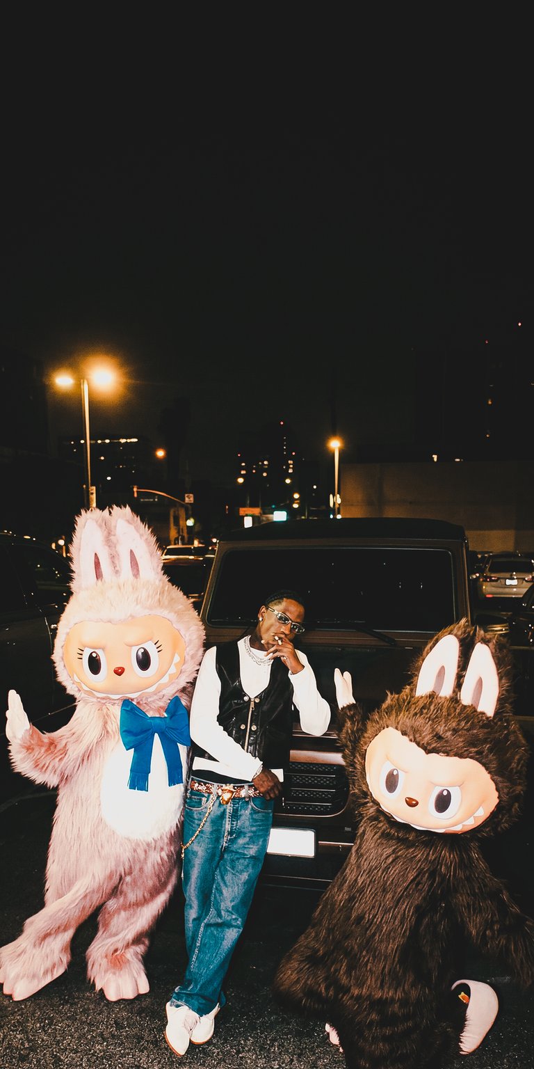 This Aesthetic wallpapers features Travis Scott posed between two large pink and brown Labubu mascots in a cinematic night setting. The 4K wallpaper for iPhone captures a moody urban vibe with warm street lighting and high-contrast shadows.