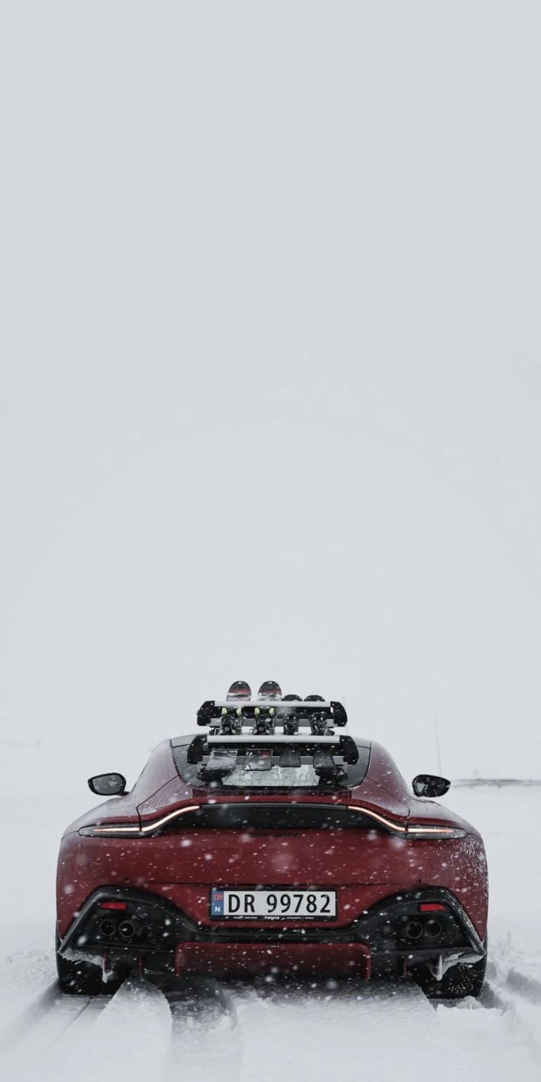 Experience the perfect blend of high-performance engineering and serene winter landscapes. This 4K wallpaper features a stunning deep red Aston Martin Vantage equipped with a roof-mounted ski rack, navigating through a crisp, minimalist snow-covered trail. The clean, white background provides a sophisticated aesthetic that highlights the sleek curves and aggressive rear profile of the vehicle, making it an ideal choice for automotive enthusiasts who appreciate a clean and modern mobile display.