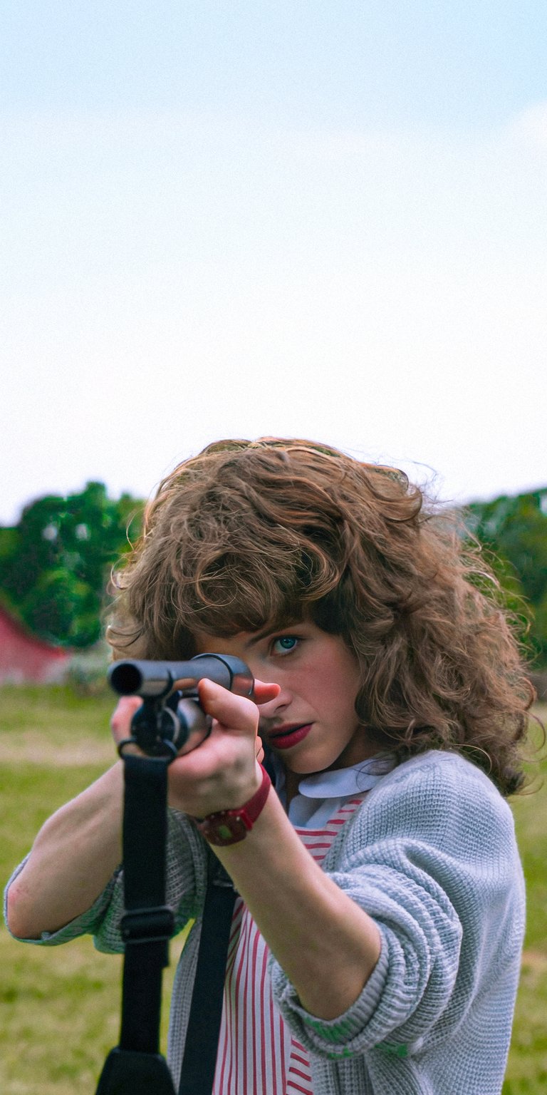 Download this incredible Nancy Wheeler 4K wallpaper for your iPhone. The detailed wallpaper features her aiming a rifle, with curly hair, and wearing a red wristwatch.