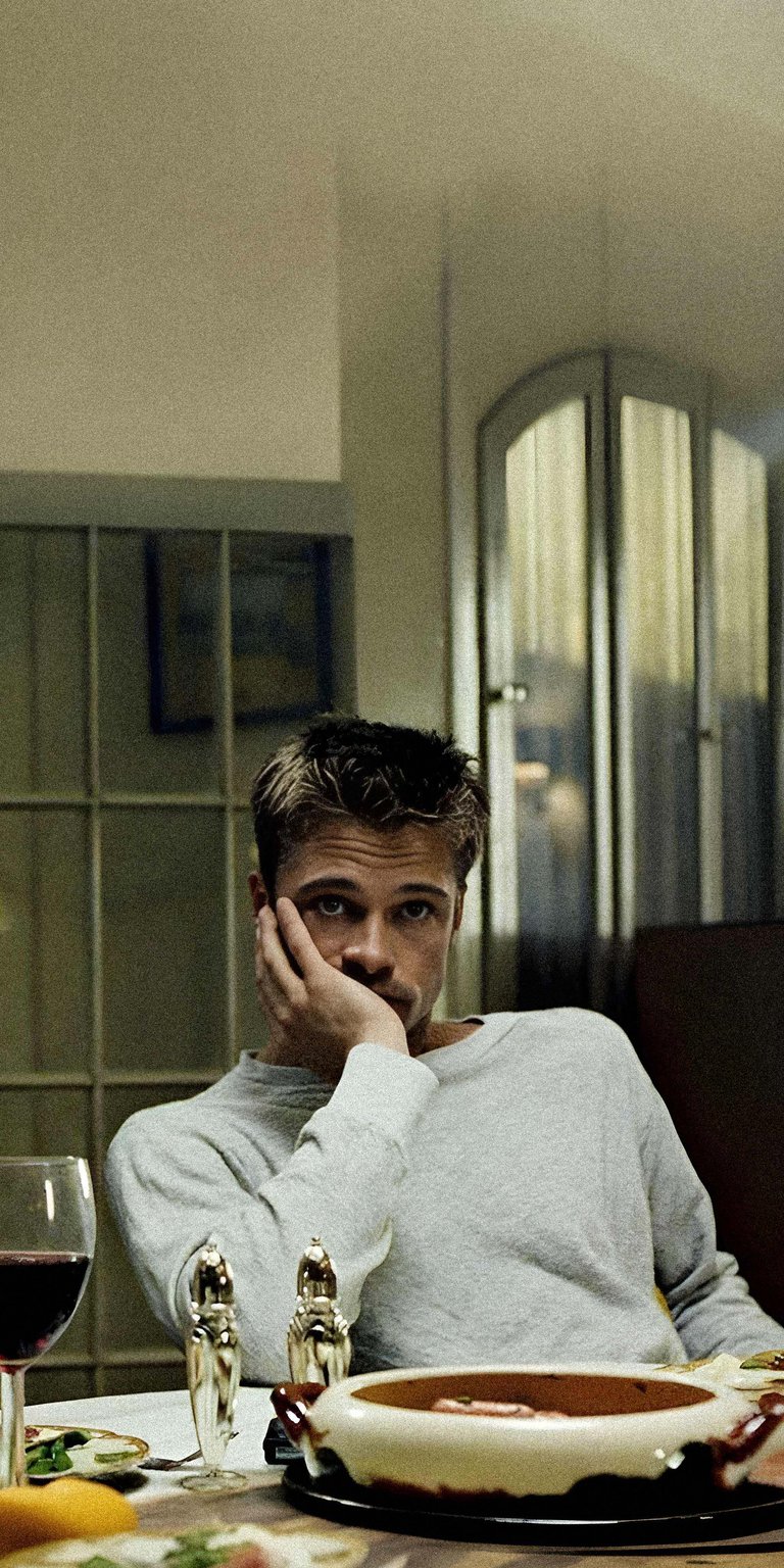 Download this Brad Pitt 4K wallpaper for your iPhone, showcasing him resting his face thoughtfully at the table. The wallpaper features a glimpse of a wine glass and decorative salt and pepper shakers.