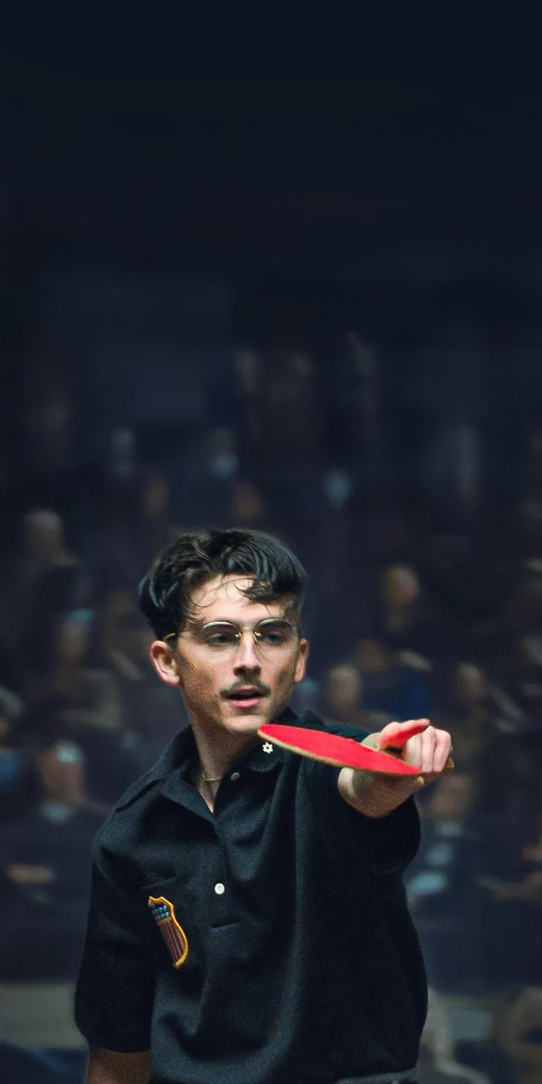 Elevate your iPhone with this striking Timothée Chalamet 4K wallpaper. The wallpaper captures Timothée in a vintage-style black polo, sporting spectacles, and holding a ping pong paddle, set against a blurred crowd.
