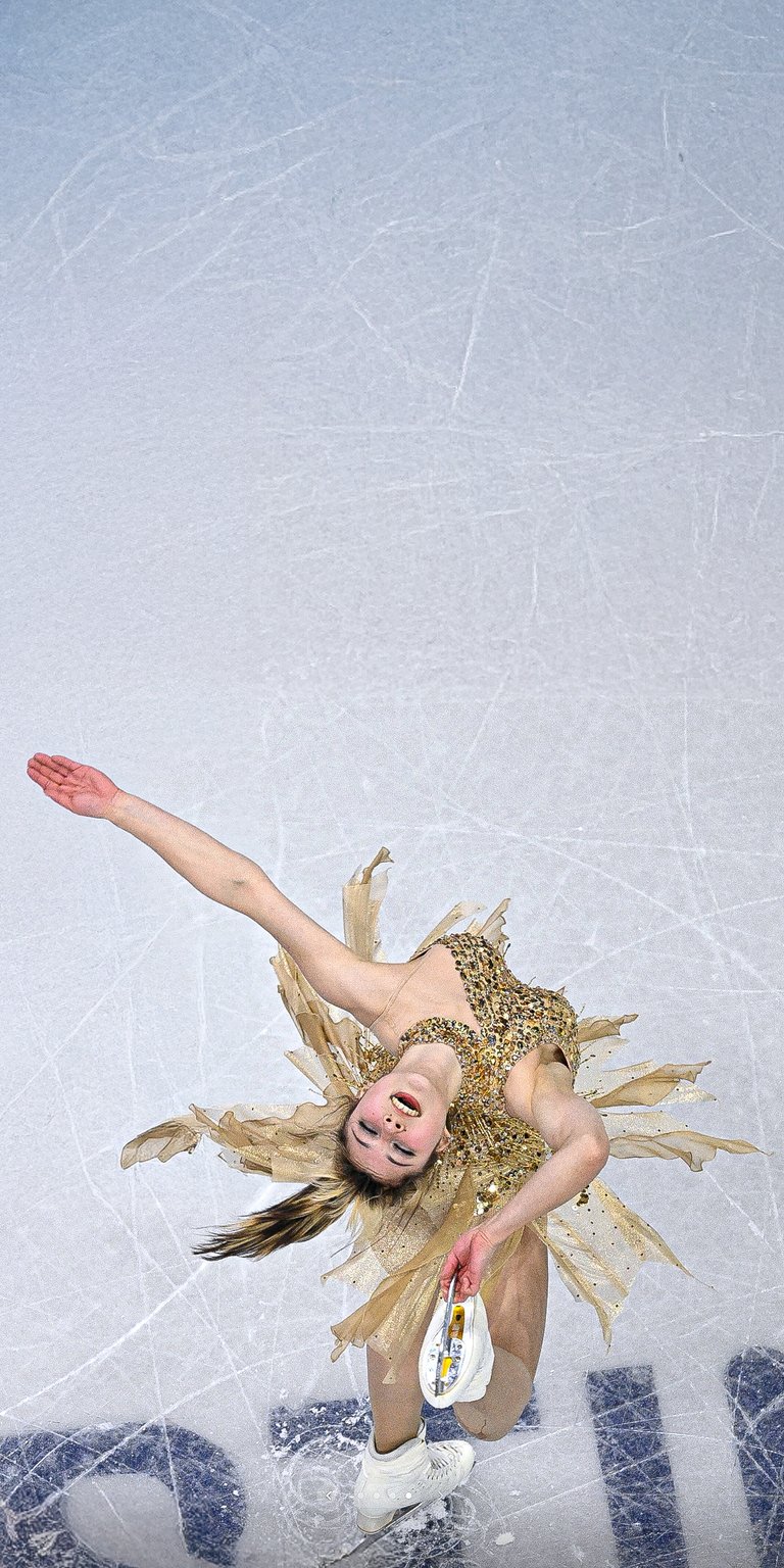 A high-angle shot of figure skater Alysa Liu performing a dynamic spin on the ice in a gold feathered dress. This sports wallpaper captures the elegance and motion of professional skating for your iPhone.