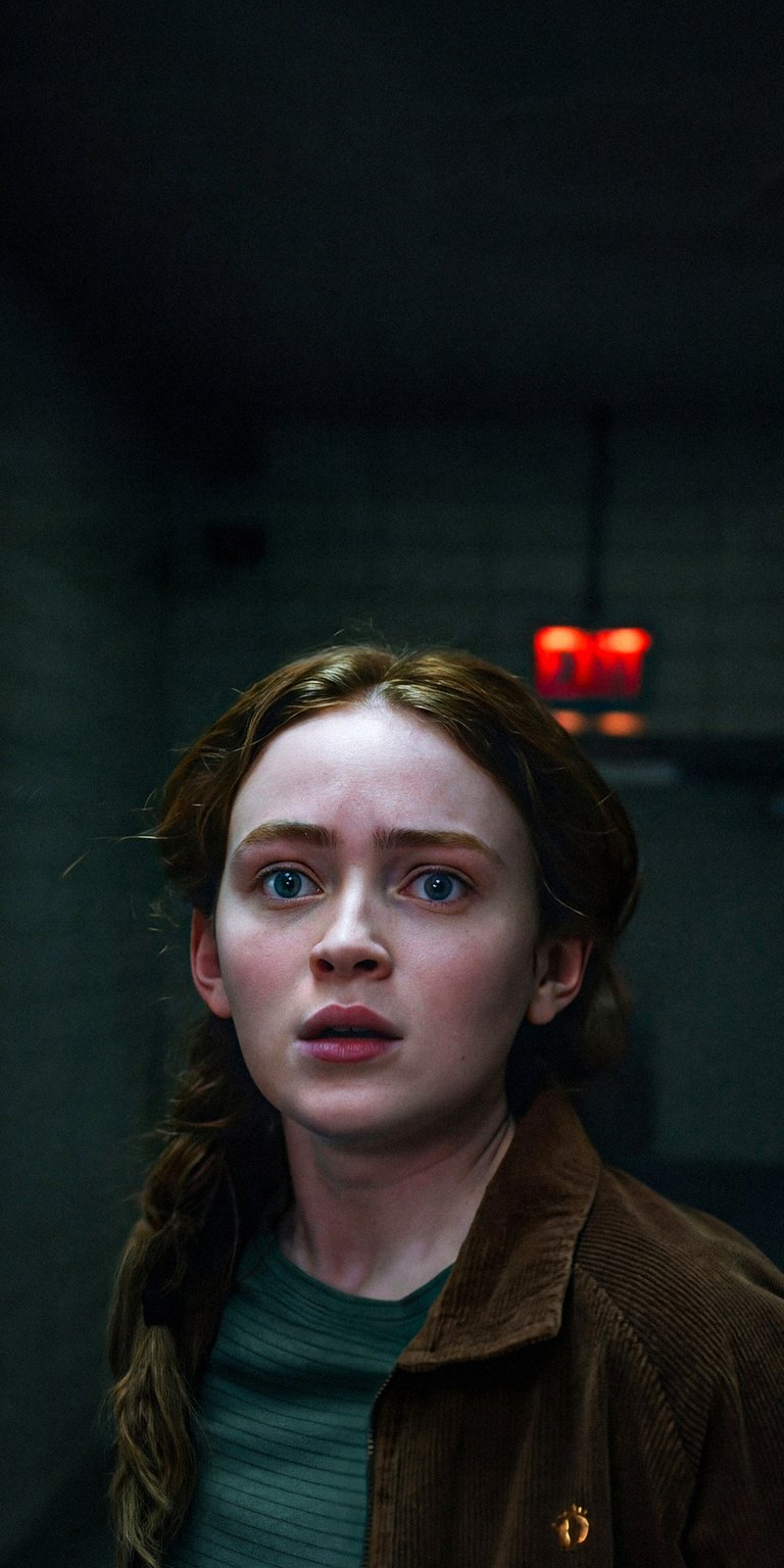 Download this captivating Sadie Sink 4K wallpaper for your iPhone, featuring a distressed expression and a partially braided hairstyle. The dark background contrasts with a red light and the golden emblem on her jacket.