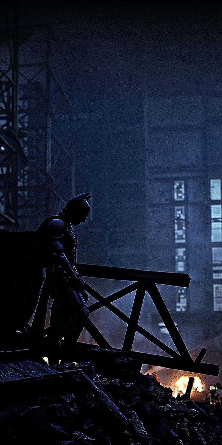 Download this Batman 4K wallpaper for your iPhone, featuring the dark knight overlooking a burning city. The high-resolution wallpaper captures Batman's silhouette against the backdrop of industrial structures and a wooden beam.