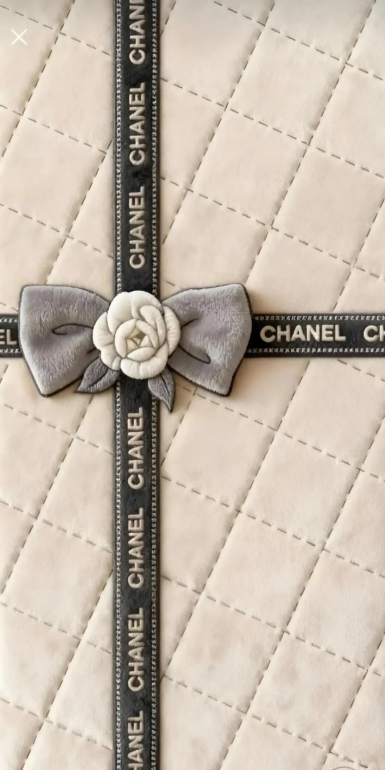 This 4K wallpaper features a luxurious cream-colored quilted texture adorned with a branded Chanel ribbon and a plush camellia flower. It is a sophisticated aesthetic wallpaper designed to bring a high-fashion, elegant look to your iPhone lock screen.
