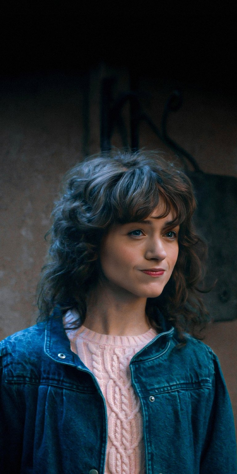 This iPhone wallpaper features Natalia Dyer in stunning 4K detail. She wears a blue jean jacket over a pink cable-knit sweater, set against a rustic, textured background, making this wallpaper a visually compelling choice.