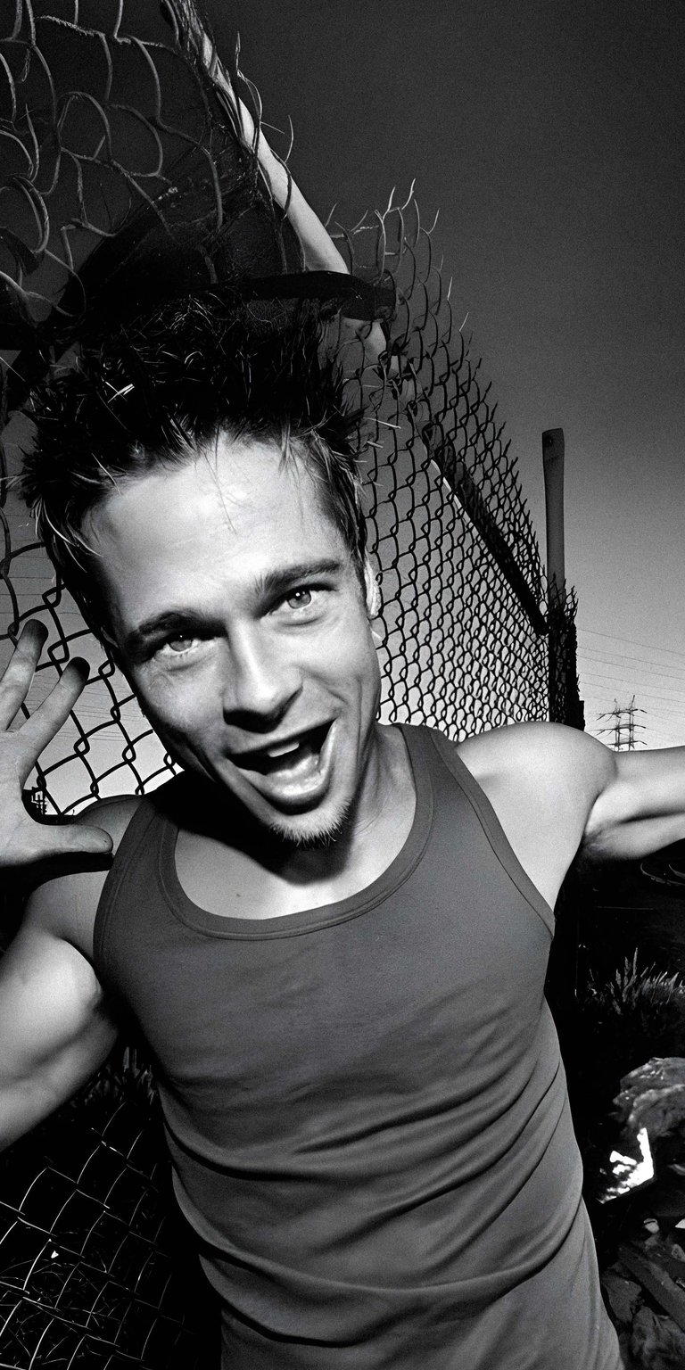 Get this Brad Pitt wallpaper for your iPhone in stunning 4K resolution, showcasing the actor leaning against a chain-link fence with an open-mouthed expression and tank top. This black and white wallpaper highlights his spiky hair and muscular physique.