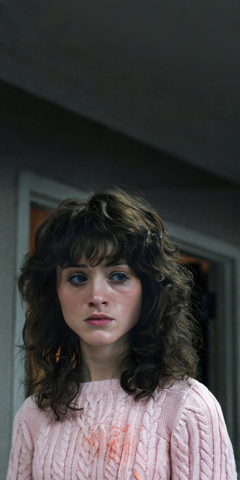 Download this incredible Natalia Dyer 4K wallpaper for your iPhone today! The wallpaper features Natalia with curly hair, wearing a pink sweater with a spot of red on it.