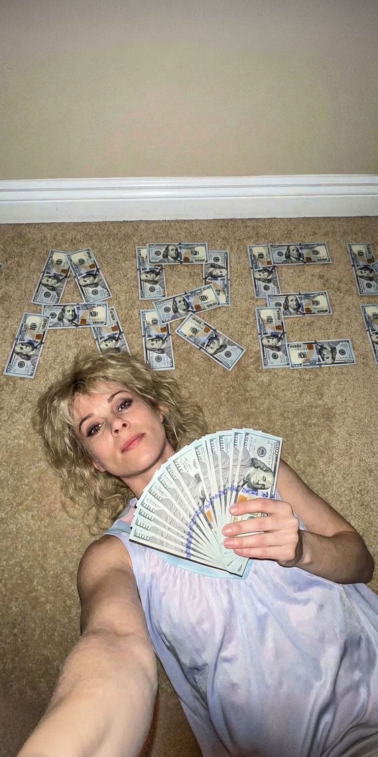 This live wallpaper features Karen Wheeler, the mother of Nancy and Mike. She is lying on the floor, surrounded by dollar bills that clearly spell out her name, "KAREN". She is dressed in a soft, light-colored nightwear or dress, capturing her classic 80s mom aesthetic. This wallpaper offers a more glamorous, domestic look from the world of Stranger Things.