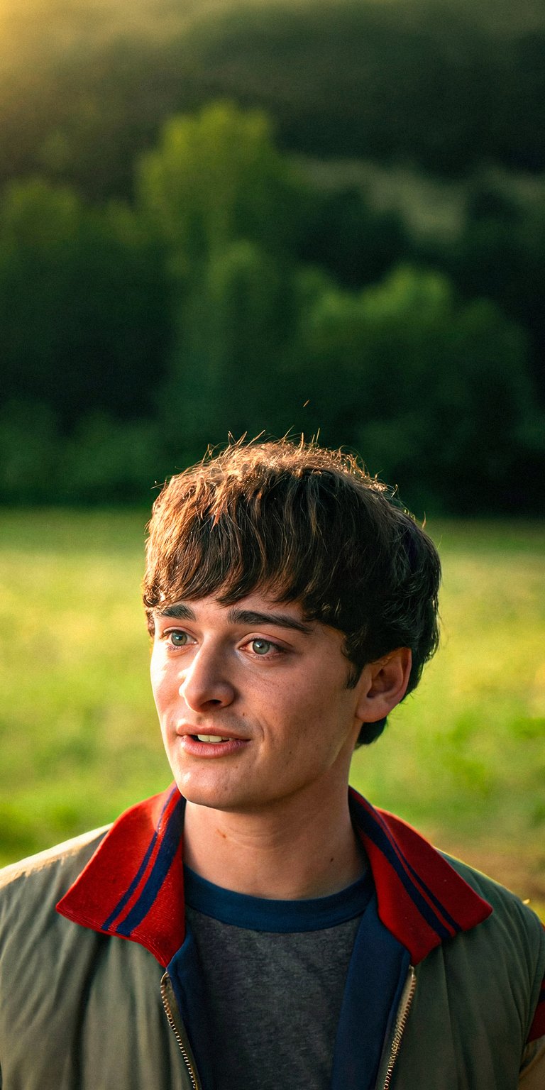 Download this Will Byers (Noah Schnapp) 4K wallpaper for your iPhone and enjoy the view of his green eyes and signature haircut. This wallpaper captures Noah in his tan jacket with a red and blue collar, set against a blurred green field.