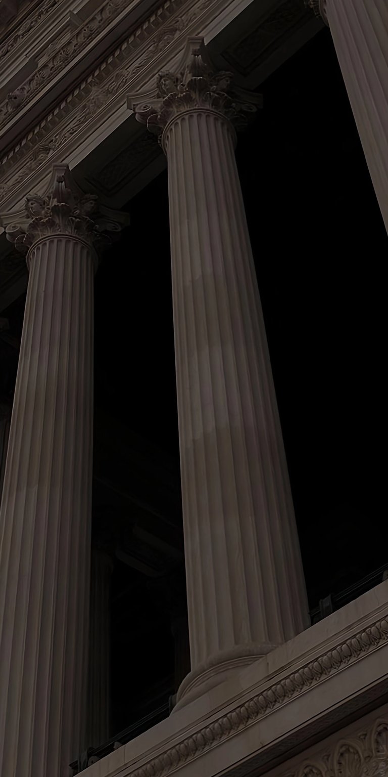 A cinematic, low-angle shot of towering Corinthian columns against a pitch-black night sky. This wallpaper captures the somber beauty of classical architecture, emphasizing the scale and texture of the stone pillars. Perfect for fans of the Dark Academia aesthetic, history, and minimalist architectural design.