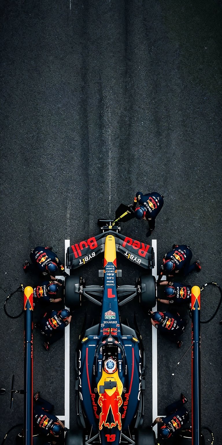 This 4K OLED wallpapers design features an aerial view of a Red Bull Racing pit stop for your iPhone. The wallpaper showcases a deep black asphalt background contrasted with vibrant navy, red, and yellow team colors.