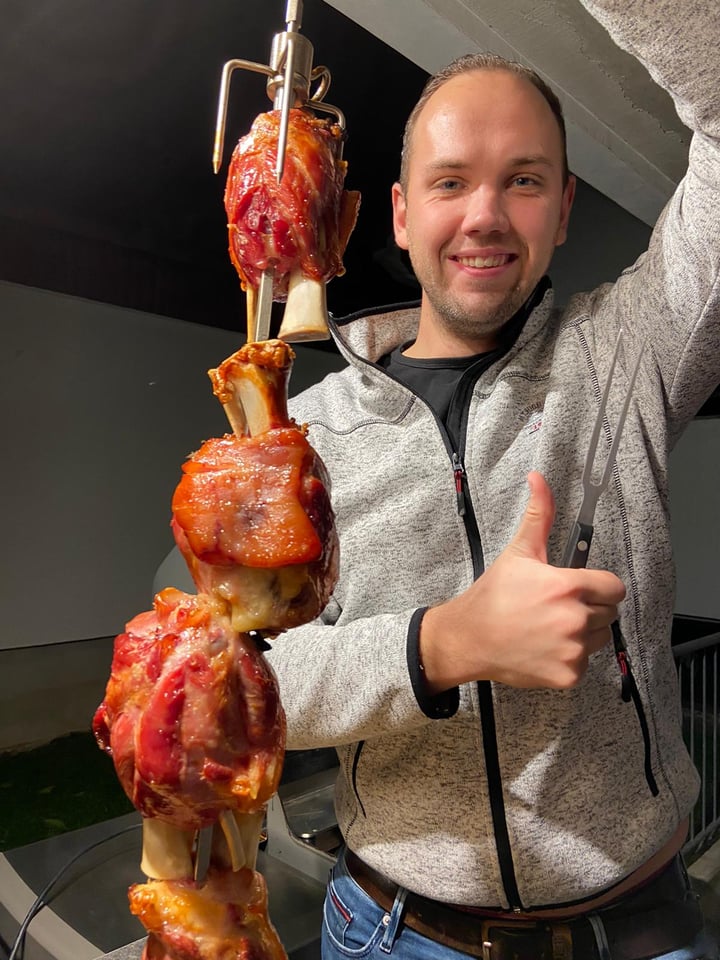 Fabian hält einen Drehspieß mit knusprigen Schweinshaxen und zeigt den Daumen nach oben