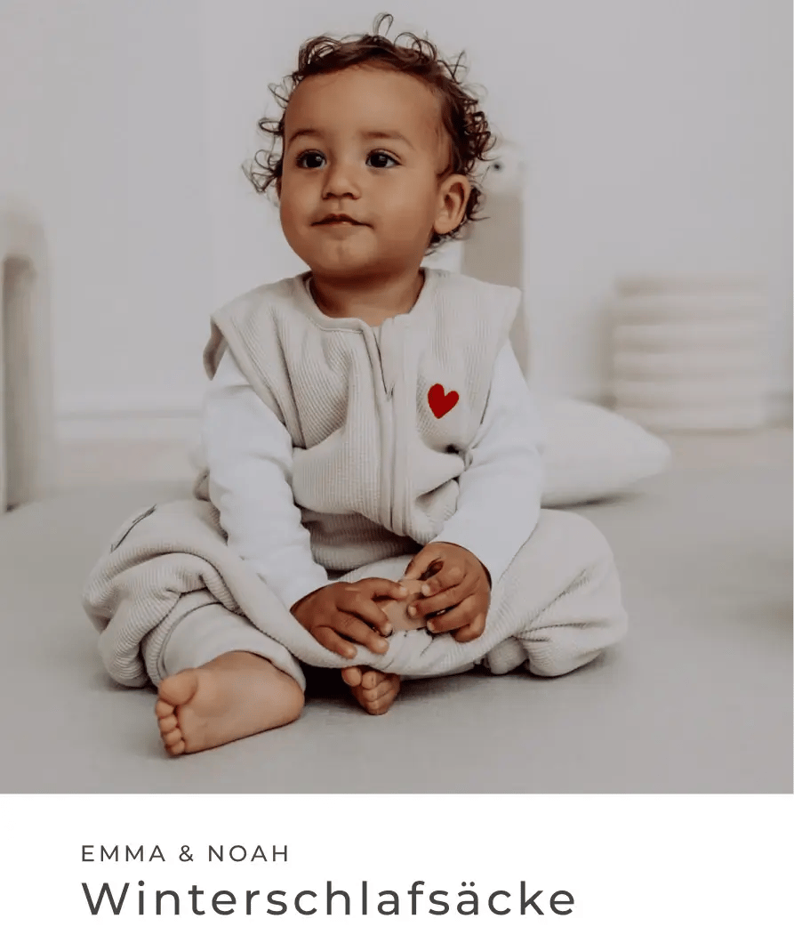 Baby sitzt in einem hellen Winterschlafsack mit rotem Herz auf der Brust vor weißem Hintergrund. Emma & Noah: Winterschlafsäcke.