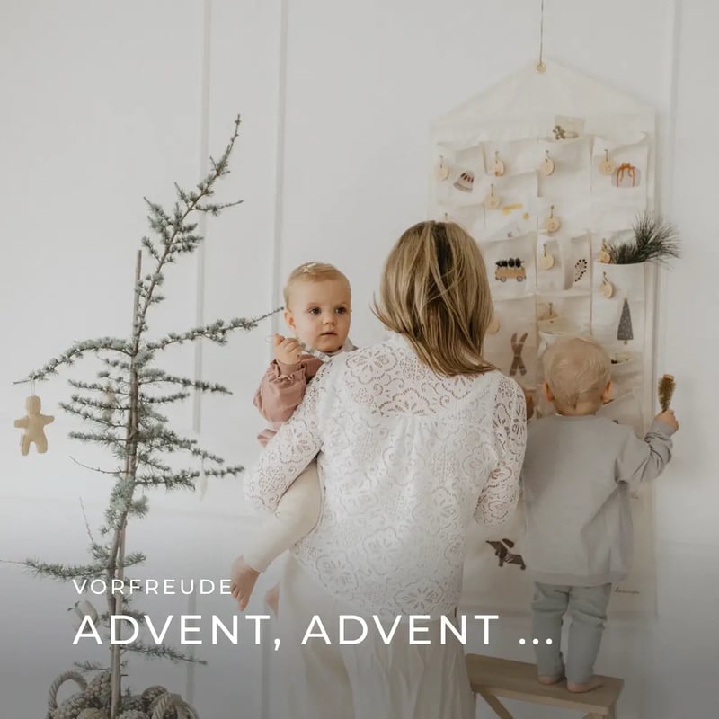 eine Mutter mit Baby auf dem Arm und ein Kleinkind auf einer Bank stehen vor einem Wand-Adventskalender