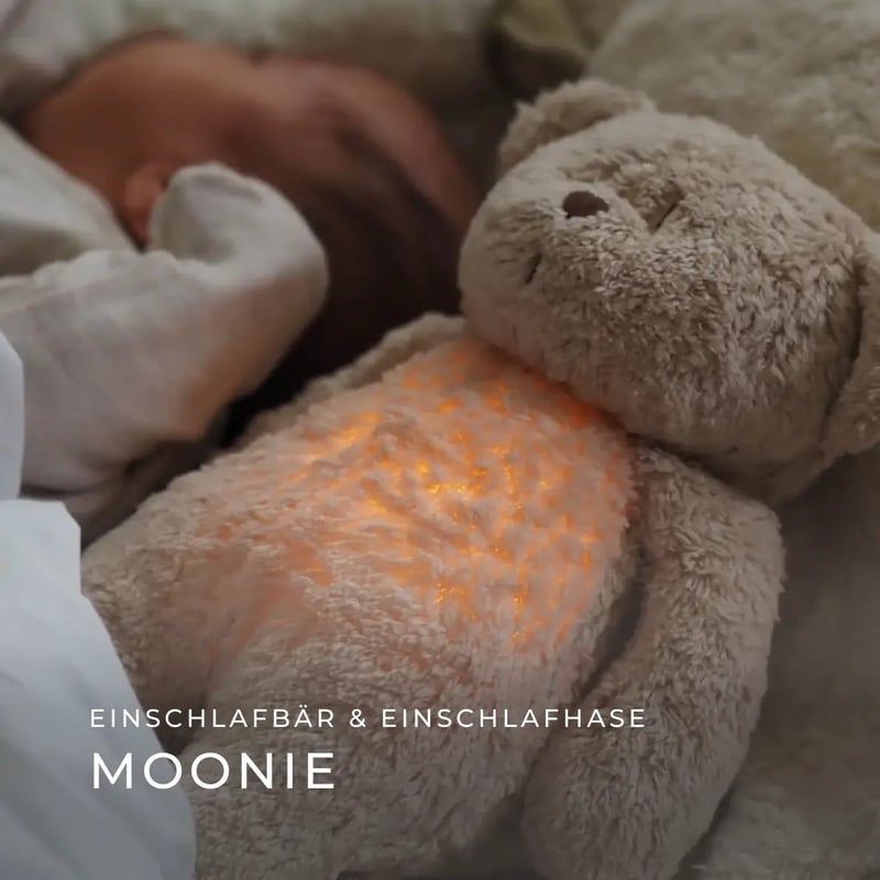 der Einschlafbär von Moonie liegt in einem Babybett, der Bauch des Bären leuchtet sanft