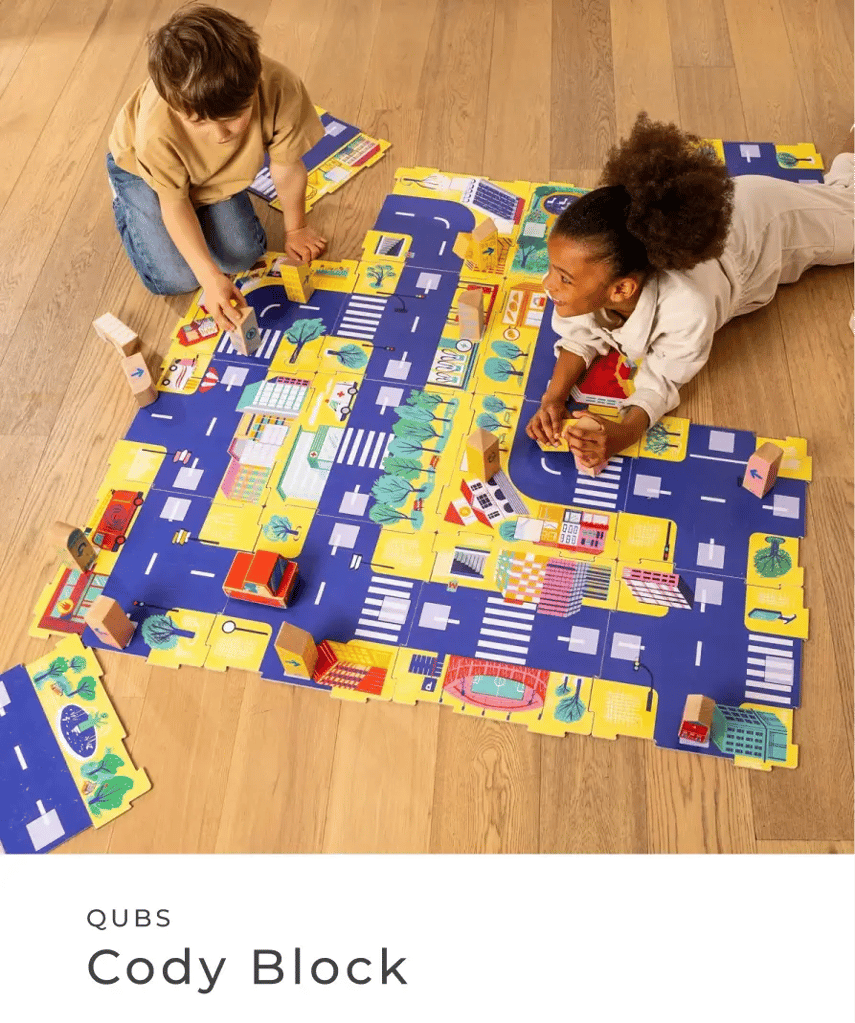 QUBS: Cody Block. Zwei Kinder spielen auf dem Boden mit einer bunten Stadt-Spielmatte und Holzbausteinen.