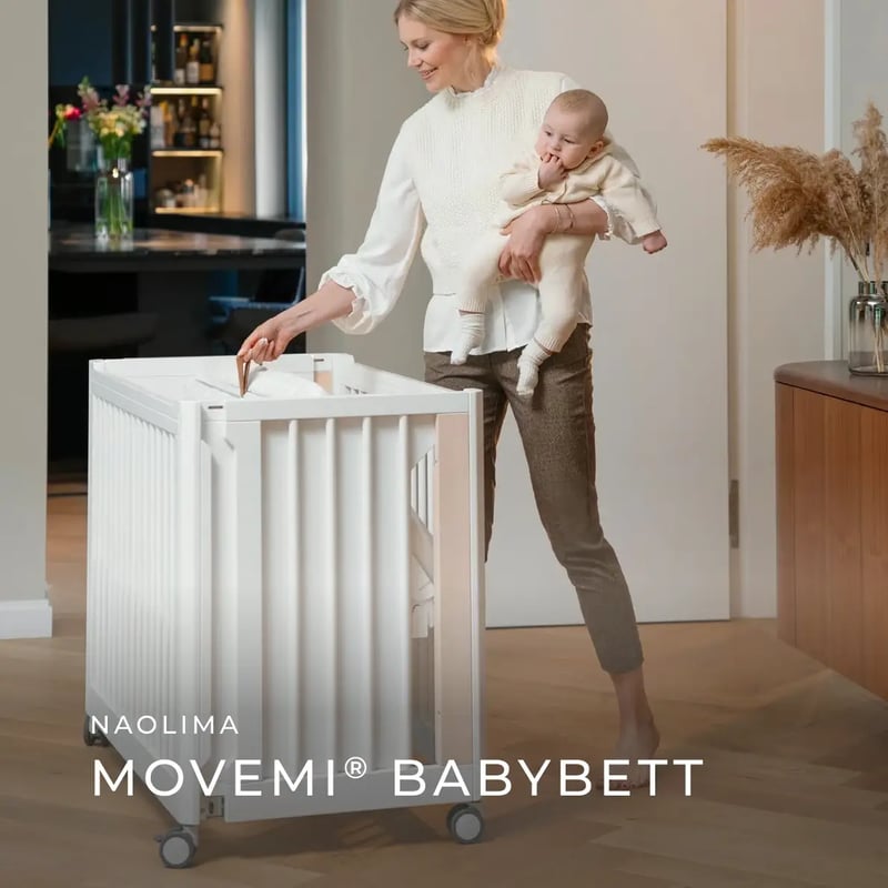 Eine Frau hält ein Baby im Arm und steht neben dem weißen, rollbaren „Movemi“ Babybett von Naolima