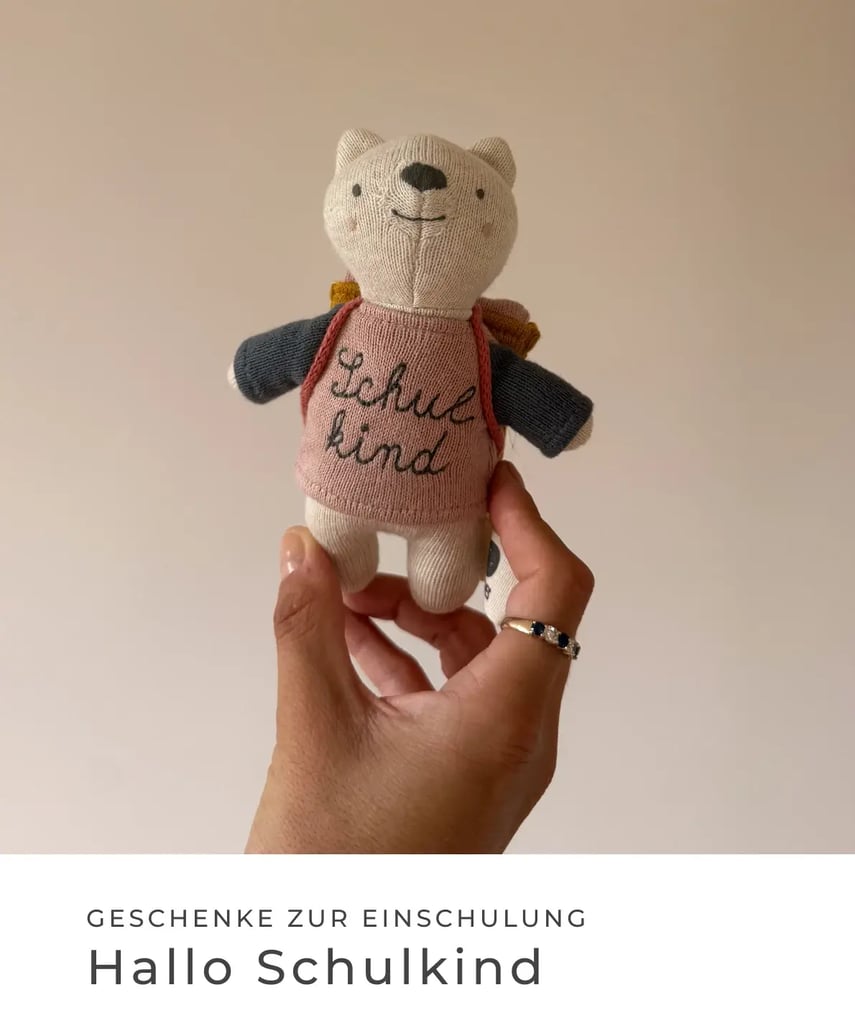 Geschenke zur Einschulung: Hallo Schulkind. Hand hält ein kleines gestricktes Stofftier mit Rucksack und der Aufschrift „Schulkind“.