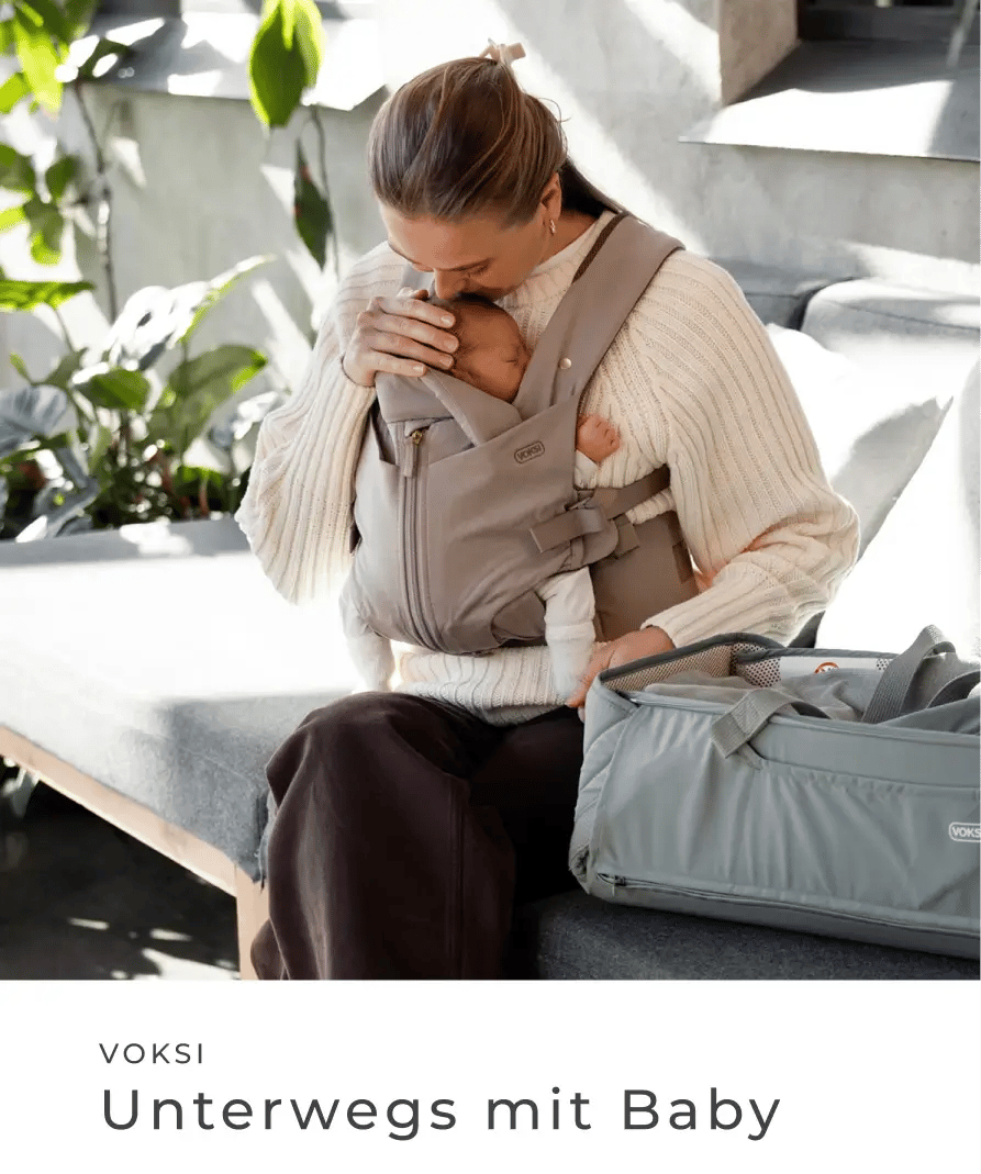 Mutter trägt ein Baby in einer Babytrage und sitzt neben einer Wickeltasche. Voksi: Unterwegs mit Baby.