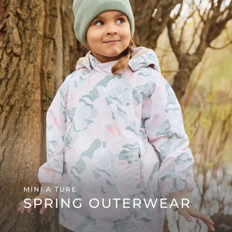 ein Kind trägt eine gemusterte, helle Übergangsjacke von Mini A Ture und steht draußen an einem Baum
