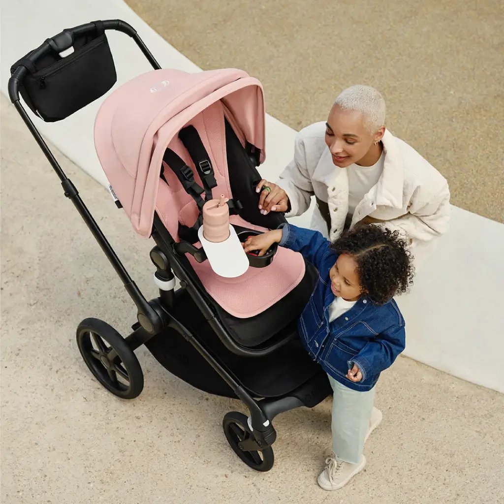 Eine Mutter mit kurzen Haaren hilft einem kleinen Kind in einem rosa Bugaboo Kinderwagen mit Becherhalter und Snacktablett, auf einer sandigen Außenfläche