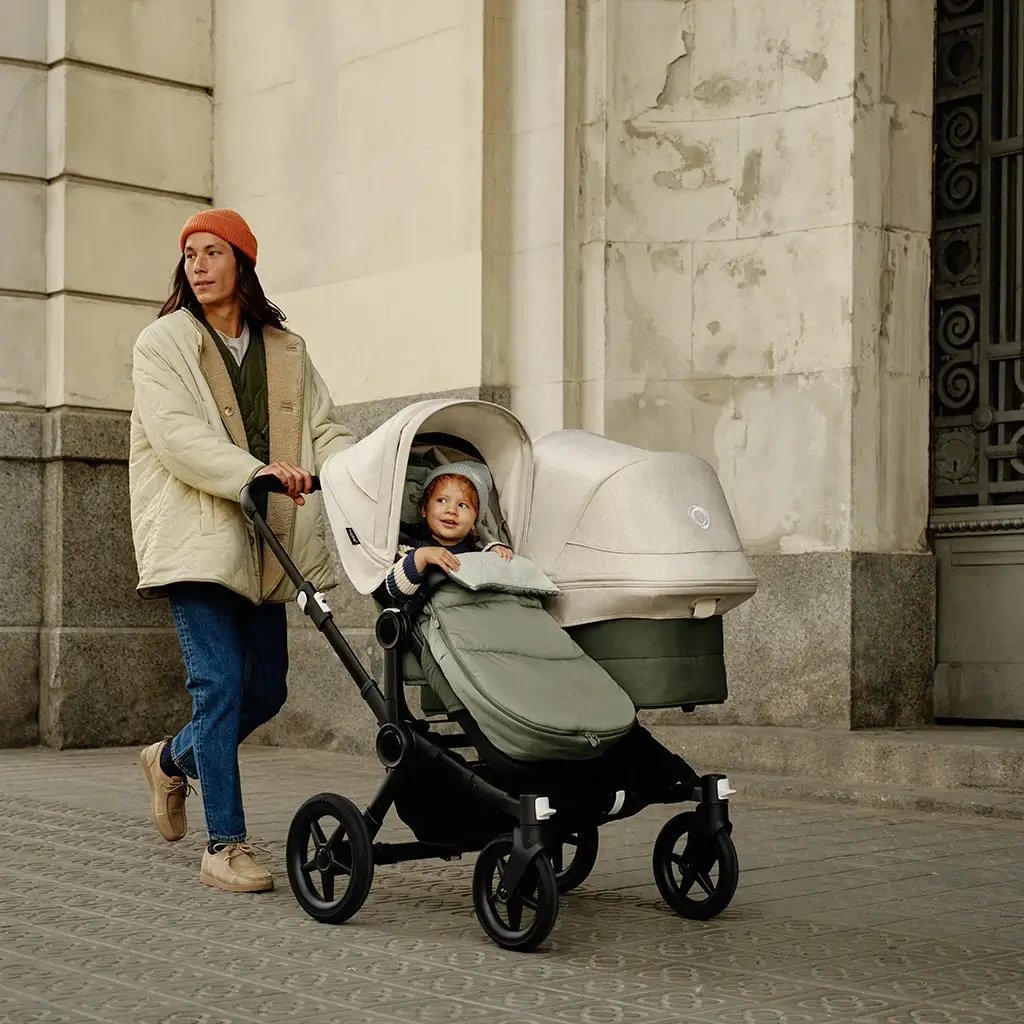 ein Vater in einer hellen Jacke und mit einer orangefarbenen Mütze schiebt einen Doppelkinderwagen von Bugaboo mit einem Kind darin, vor einem historischen Steingebäude