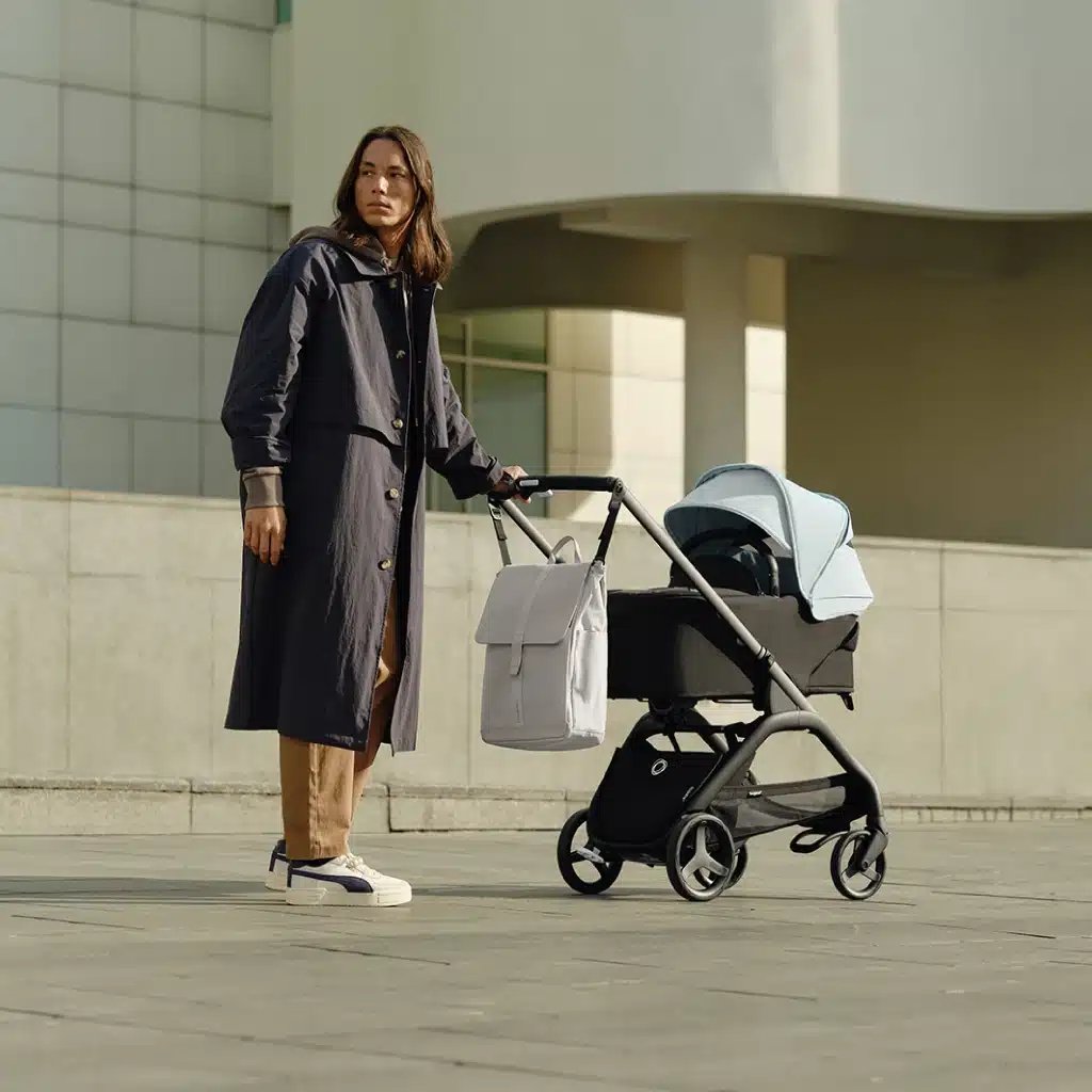 in Vater in einem langen Mantel und Turnschuhen steht neben einem Bugaboo Kinderwagen mit einem hellblauen Verdeck und einem grauen Rucksack, vor einem modernen Betongebäude