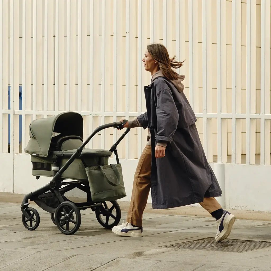 ein Vater in einem langen Mantel und Turnschuhen schiebt einen olivgrünen Bugaboo Kinderwagen auf einer Stadtstraße, mit einem großen weißen Gebäude im Hintergrund