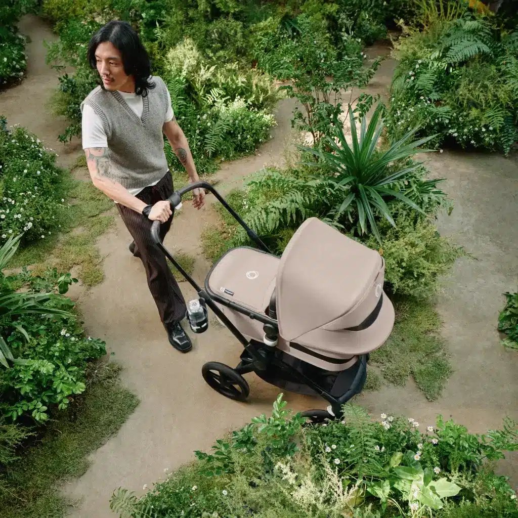 ein Vater schiebt einen beigen Bugaboo Kinderwagen durch einen grünen Garten mit üppigen Pflanzen auf beiden Seiten des Weges