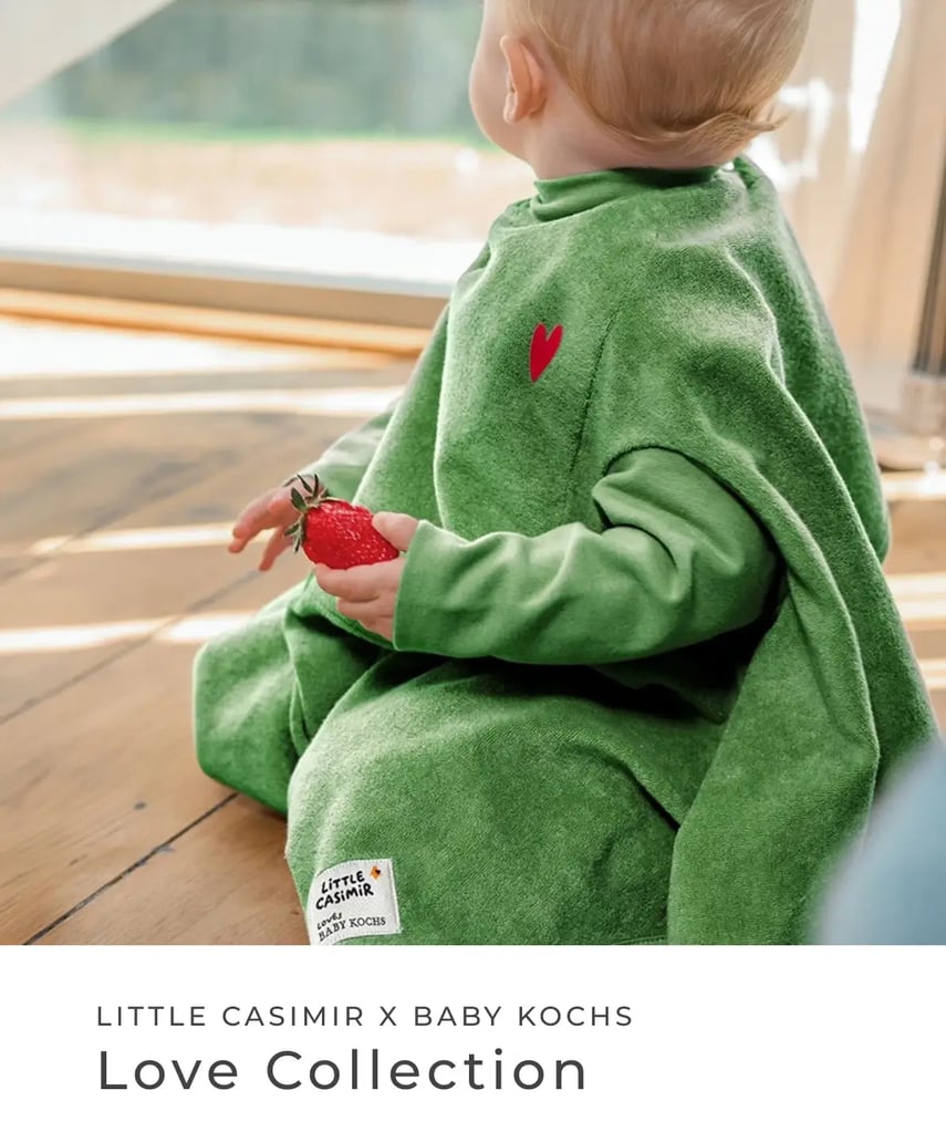 Little Casimir x Baby Kochs: Love Collection. Kleinkind sitzt in grünem Lätzchen mit rotem Herz auf dem Holzboden und hält eine Erdbeere in der Hand.
