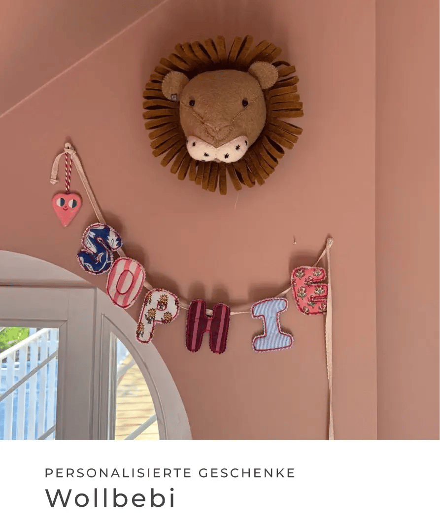 Personalisierte Namensgirlande mit dem Namen Sophie hängt an einer rosa Wand unter einem Löwenkopf. Personalisierte Geschenke: Wollbebi.