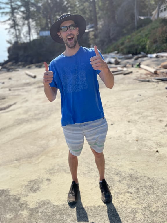 Cole wearing a blue shirt, striped shorts, and sunglasses, smiling and giving thumbs up on a sunny beach