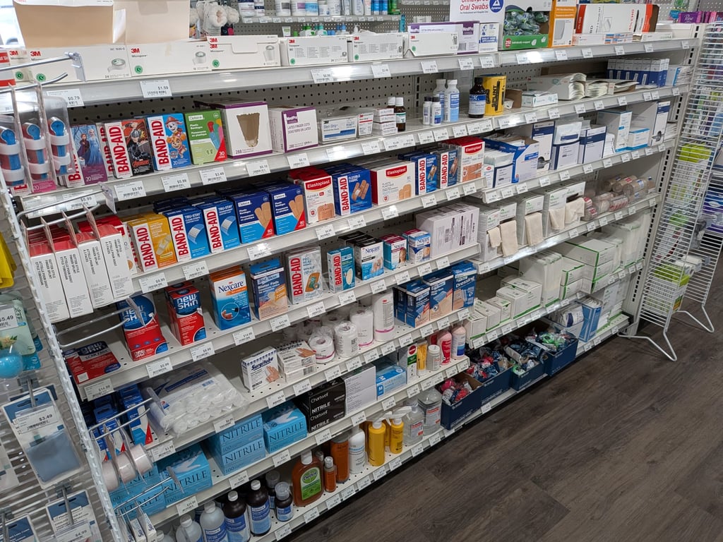 Pharmacy shelf stocked with various first aid supplies, including Band-Aids, adhesive bandages, antiseptic creams, gauze pads, nitrile gloves, medical tapes, and other health products. The shelves are organized with neatly labeled sections, displaying a wide range of healthcare items.