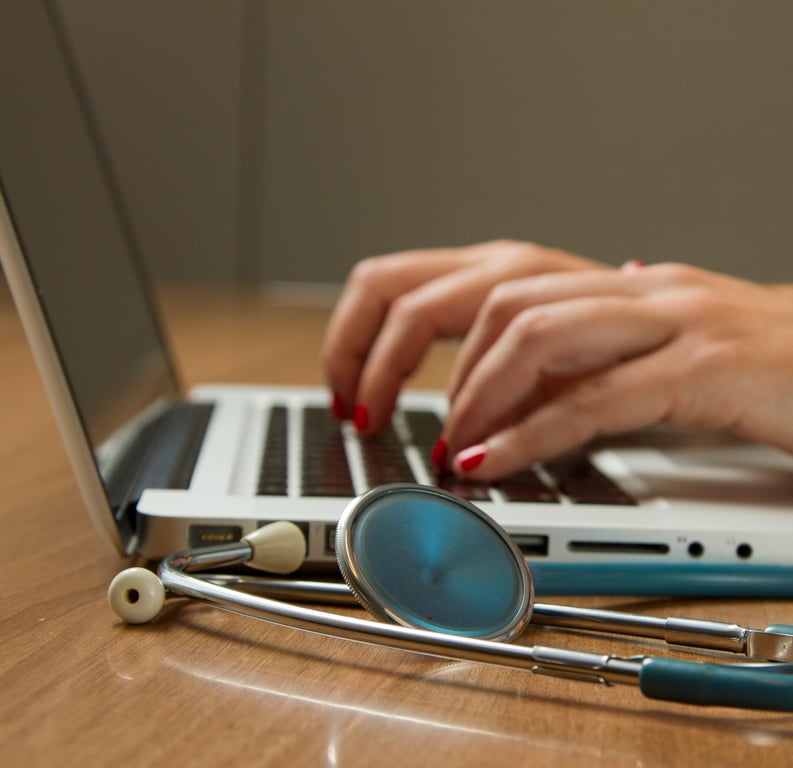 Close-up of hands typing on a laptop keyboard with a stethoscope resting on the table, symbolizing medical research or healthcare technology.