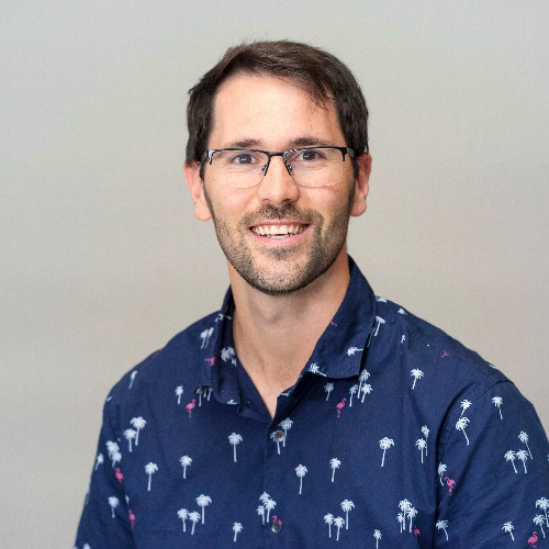 Cole wearing glasses and a navy shirt with palm tree patterns, smiling against a neutral background