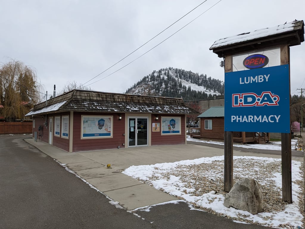 Exterior of Lumby I.D.A. Pharmacy on a snowy day with a blue signboard reading 'Lumby I.D.A. Pharmacy' and a small building with red siding and large windows.