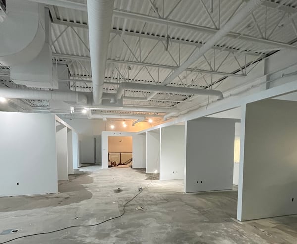 Spacious commercial interior under construction, with drywall partitions forming rooms, exposed white ceiling ducts and beams, and a concrete floor, illuminated by both natural and artificial light.
