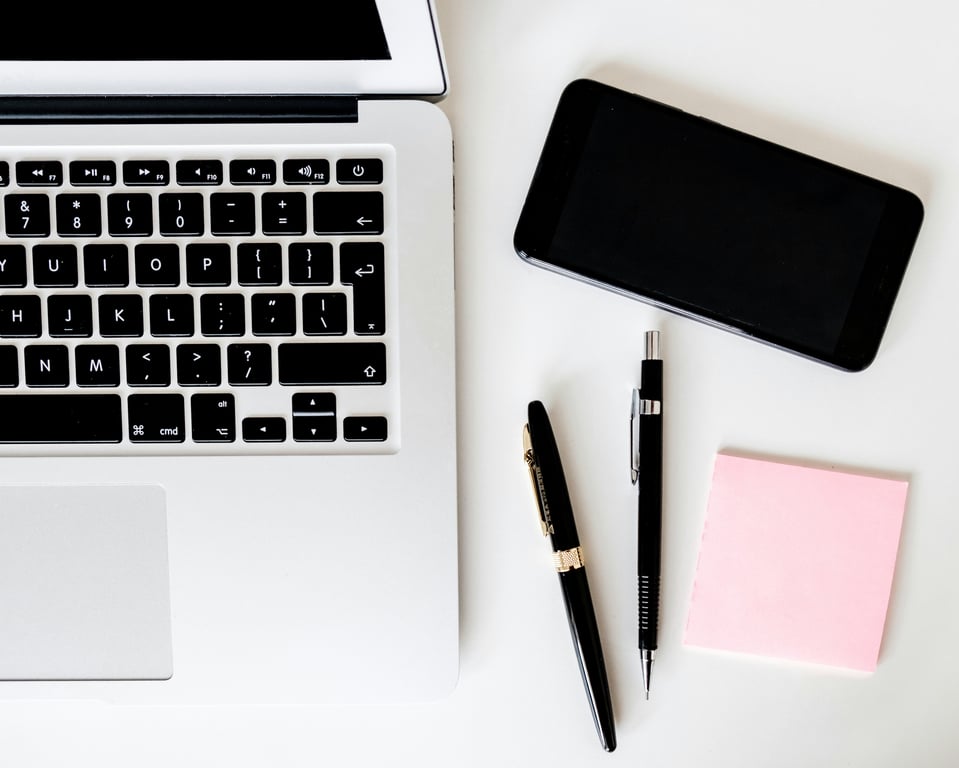 Top view of a laptop keyboard, smartphone, pens, and pink sticky notes arranged on a white desk