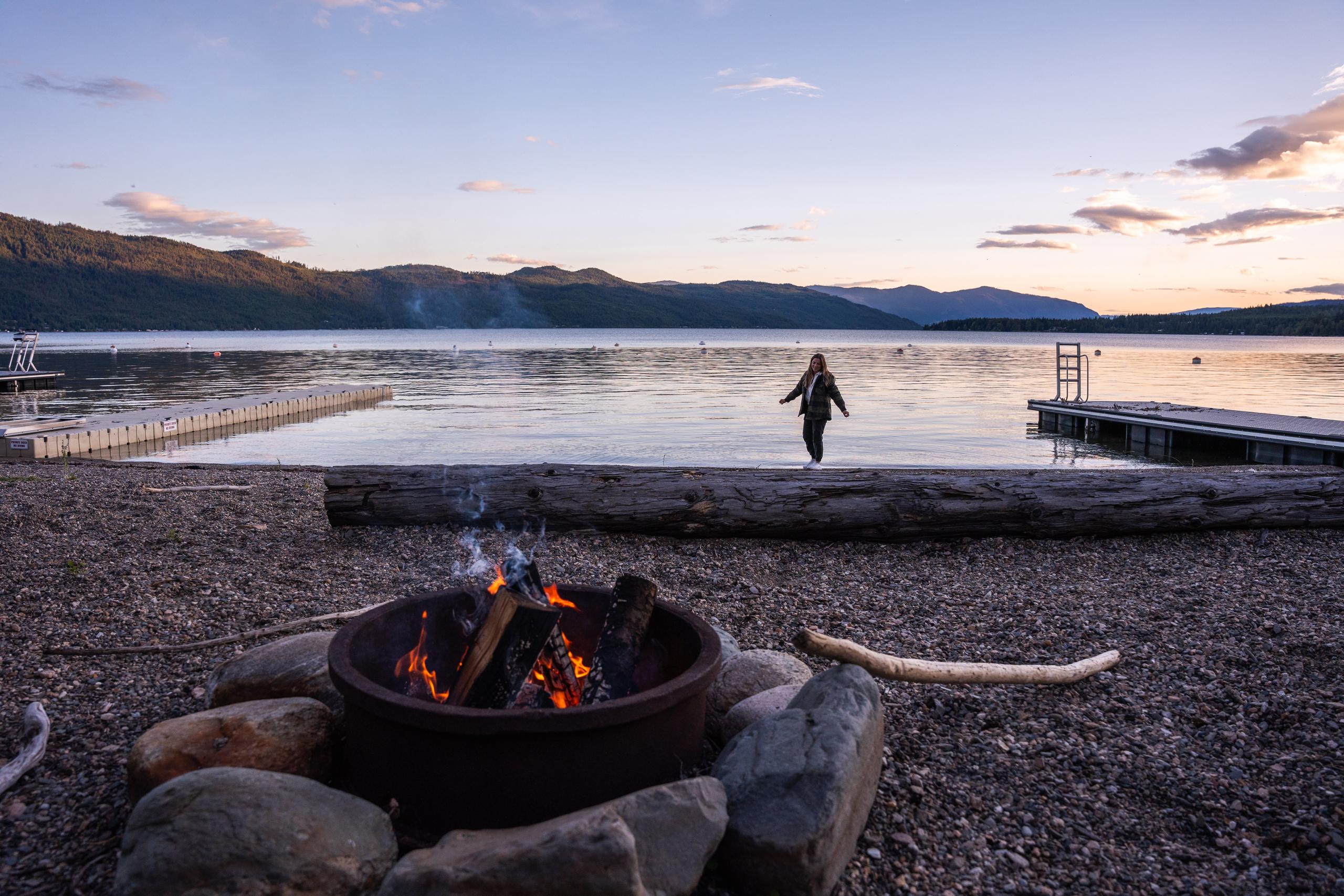 A fire ring burning on the pebble beach at sunset, with the swim dock and the lake stretching to the hills.